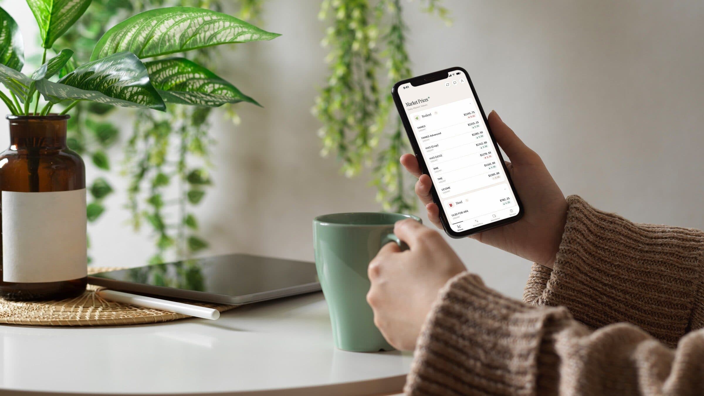 Uma mão segura um telemóvel aberto no ecrã de preços diários da app Bardi Fuels, ao lado de plantas e de uma caneca