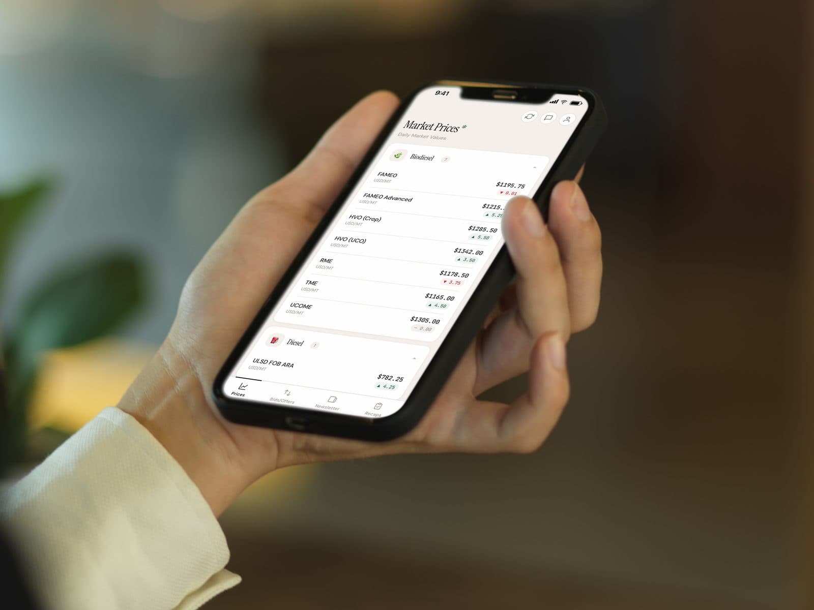 A hand holding a phone in a dim room, showing the market prices screen of the Bardi Fuels iOS app