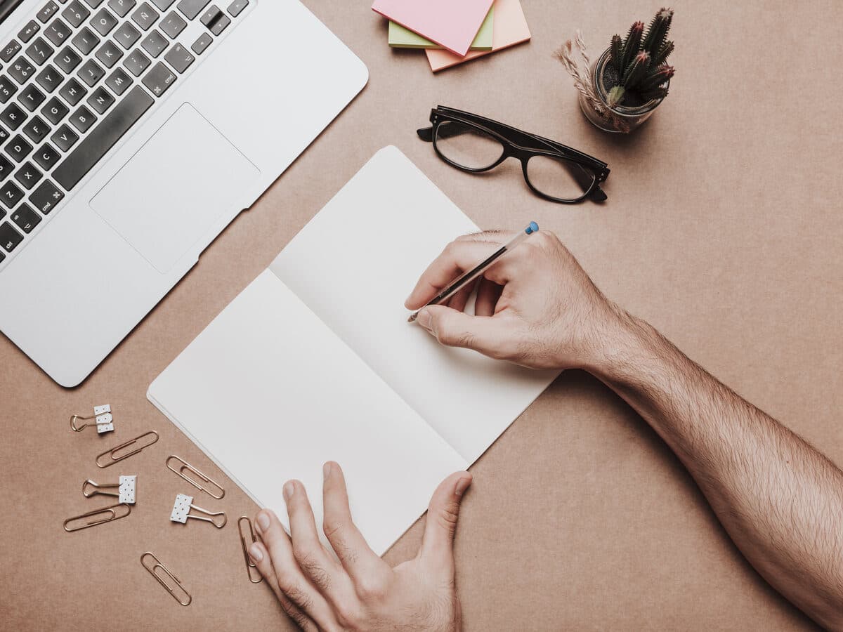 Mãos a tomar notas num caderno ao lado de um portátil