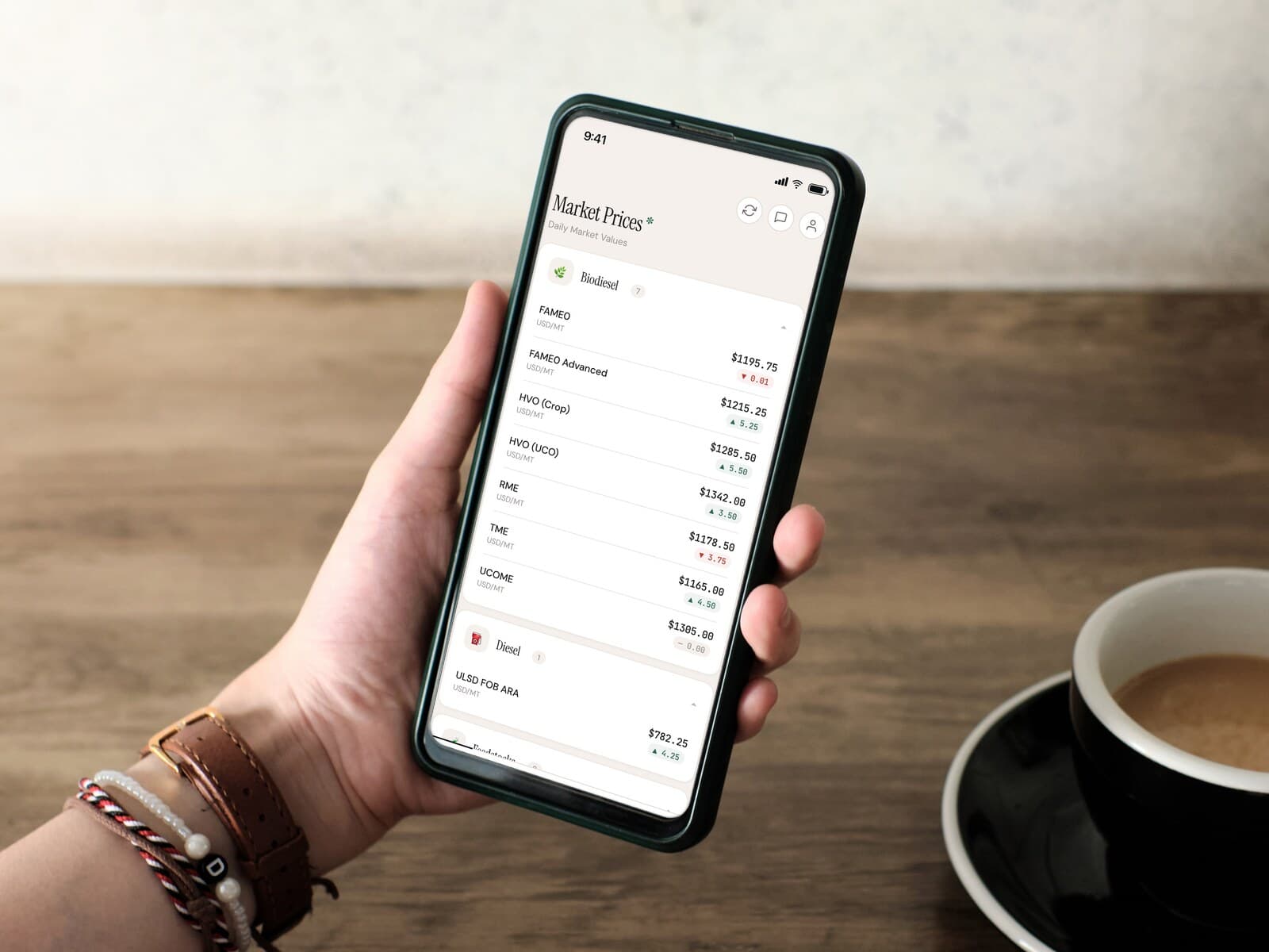 A hand holding a phone showing the Bardi Fuels daily market prices, on a wooden table beside a cup of coffee